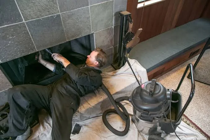 Technician cleaning the inside of a tiled fireplace beside a canister vacuum.