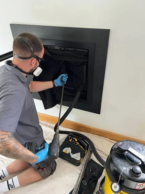 Technician in protective gear cleaning a fireplace opening with a rod and vacuum equipment.