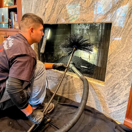 Person cleaning soot from an open fireplace with a chimney brush and vacuum hose.